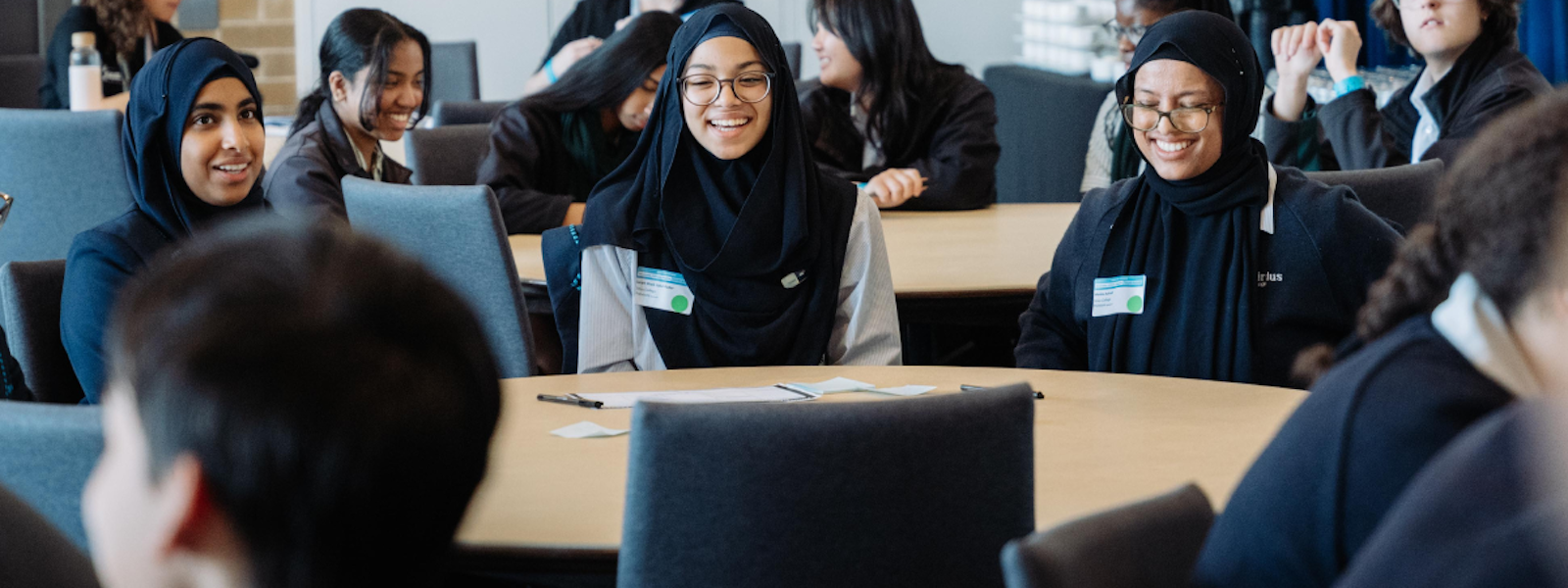 Students sit at round tables in a classroom, smiling and engaged in discussion during a collaborative learning or workshop session.